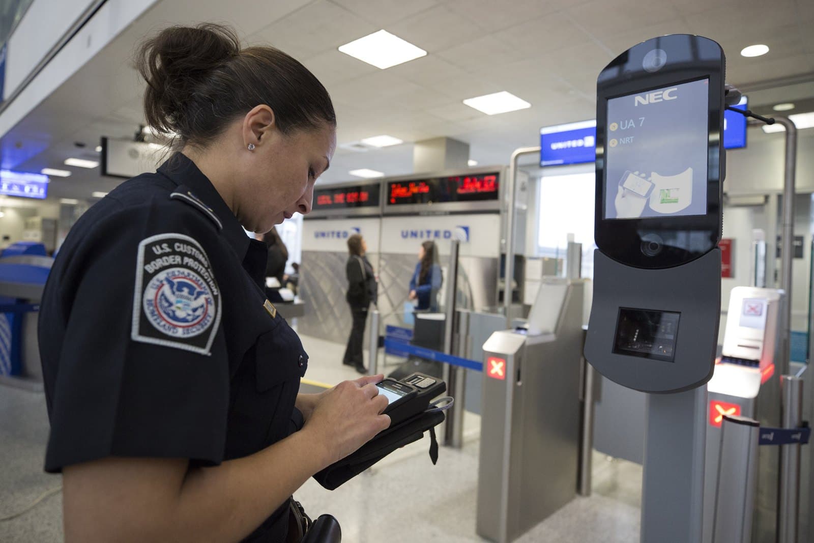 A uniformed U.S. Customs and Border Protection officer checks a handheld device next to a biometric boarding camera kiosk at an airport departure gate in Houston. Archive photo by CBP Photography, public domain, via Wikimedia Commons.