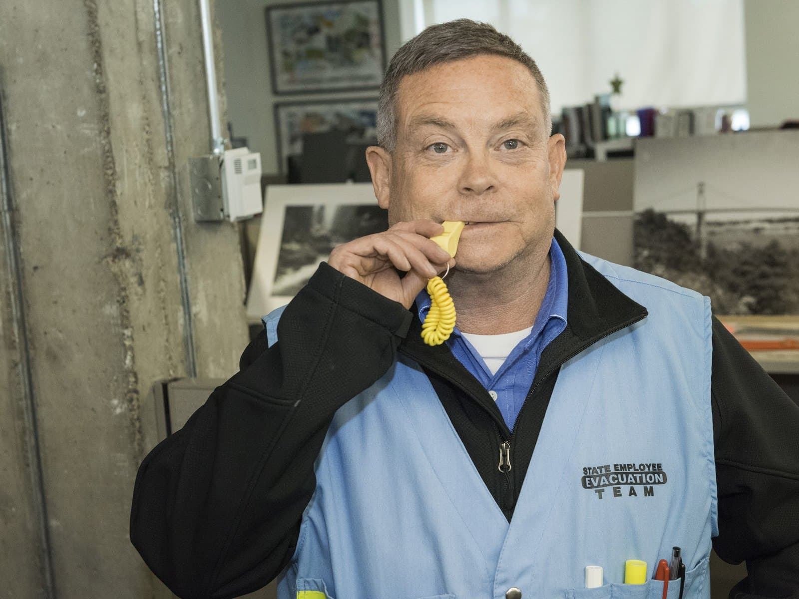 An Oregon Department of Transportation employee in a blue safety vest blows a whistle in an office to start the Great ShakeOut earthquake drill on Oct. 17, 2019. Photo by the Oregon Department of Transportation, CC BY 2.0, via Wikimedia Commons.