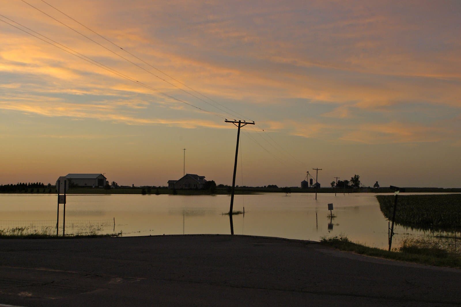 Floodwater covers rural Indiana farmland and roads at sunset; farm buildings and utility poles are silhouetted against an orange sky reflected in standing water. File photo, June 13, 2004.