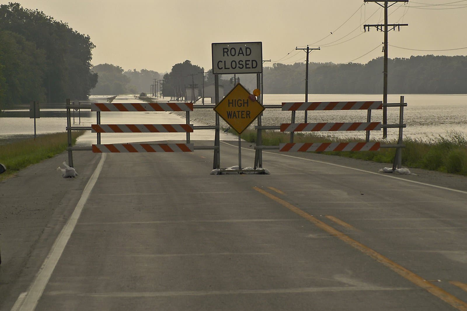 Road Closed and High Water signs barricade a flooded two-lane highway in Indiana; standing floodwater covers the roadway beyond the barricades under an overcast sky. File photo, June 13, 2004.