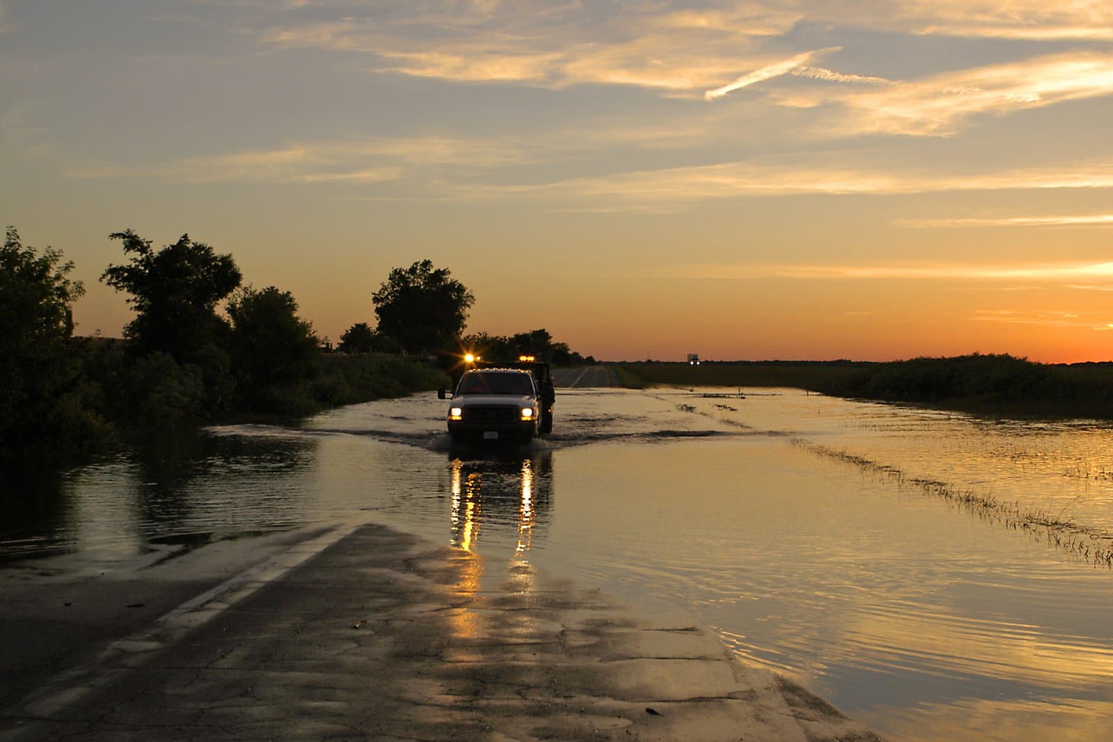 A county road truck drives through deep floodwater covering Indiana Highway 52 at sunset; headlights and amber roof lights reflect on the water. File photo, June 15, 2004.
