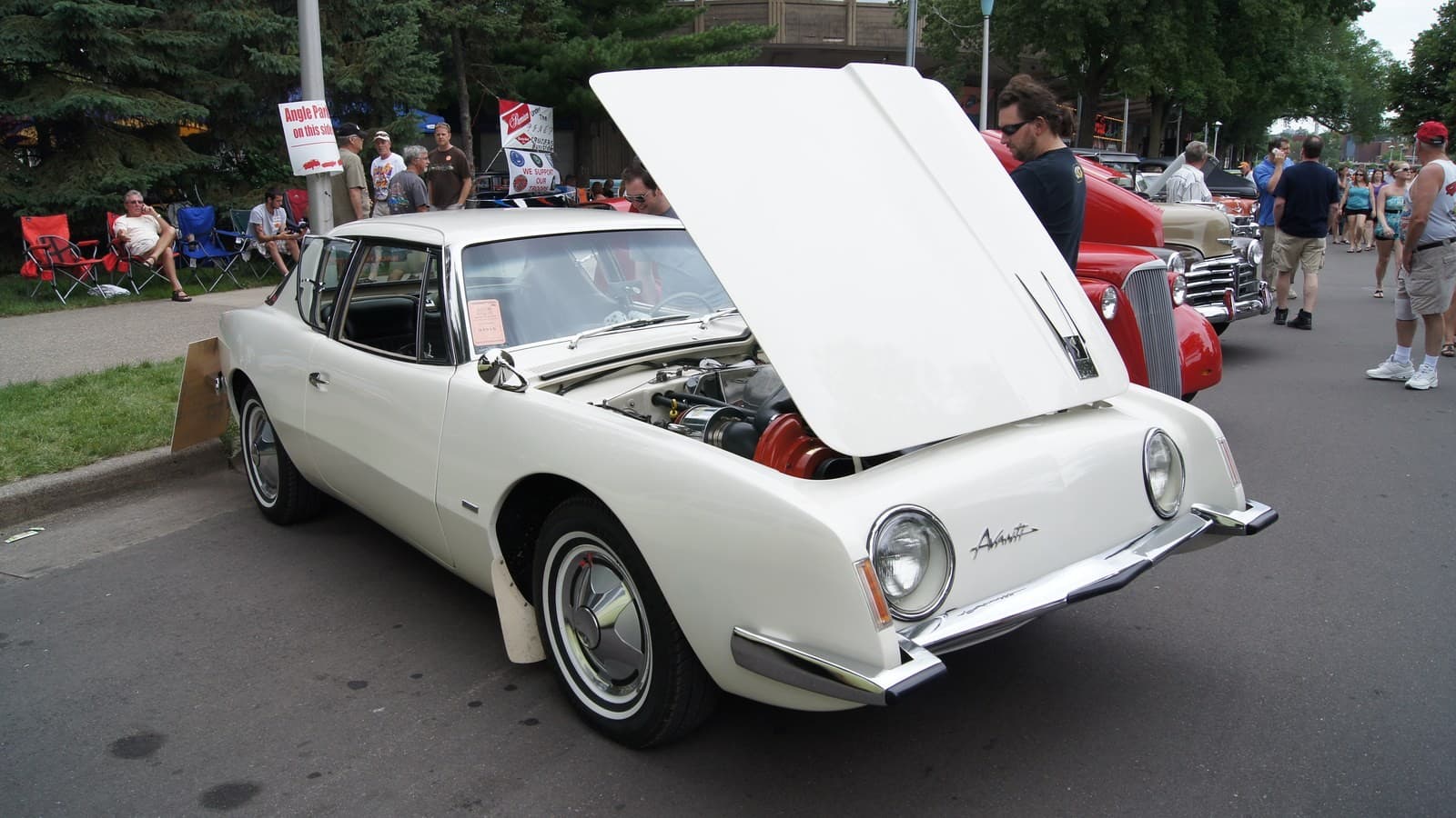 A white 1963 Studebaker Avanti parked on a street at an outdoor car show with its long hood raised, showing a red-and-chrome supercharged V8, as people stand and sit in lawn chairs nearby.